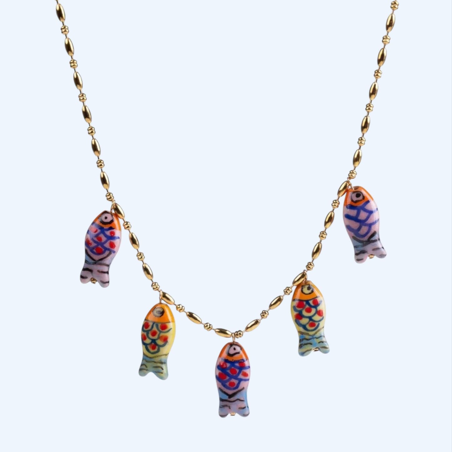 Vista frontal do Colar dourado em aço inoxidável e pendentes em forma de peixe coloridos em porcelana. Este colar tem um acabamento artesanal e vibe mediterrânica, é um colar divertido, leve, descontraído e um toque summer, beachy e artsy. Colar ideal para férias.