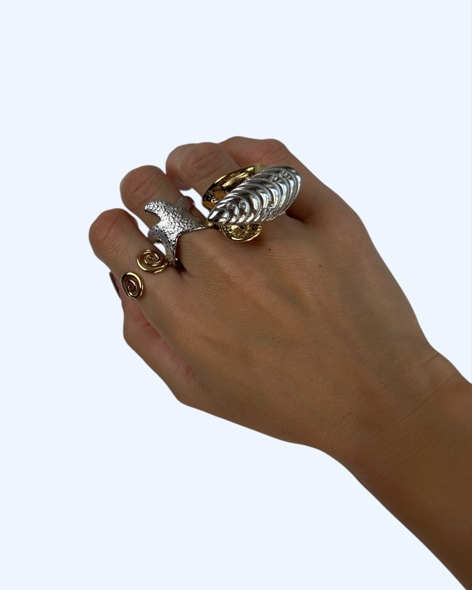 Vista a ser usado na mão, do anel metalizado com concha em aço inoxidável. É um anel bold e texturizado dourado com uma concha prateada no topo.