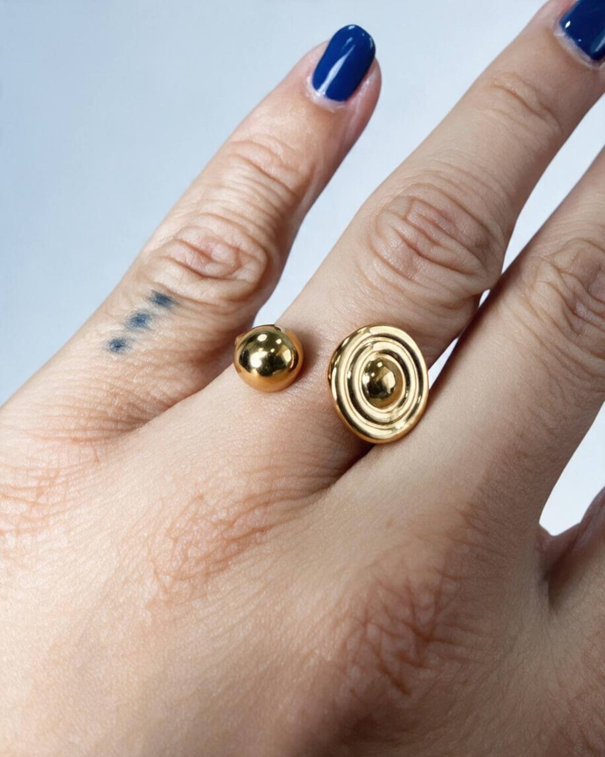 Pormenor do anel Órbita dourado no dedo, com disco de círculos concêntricos e esfera polida nas extremidades.