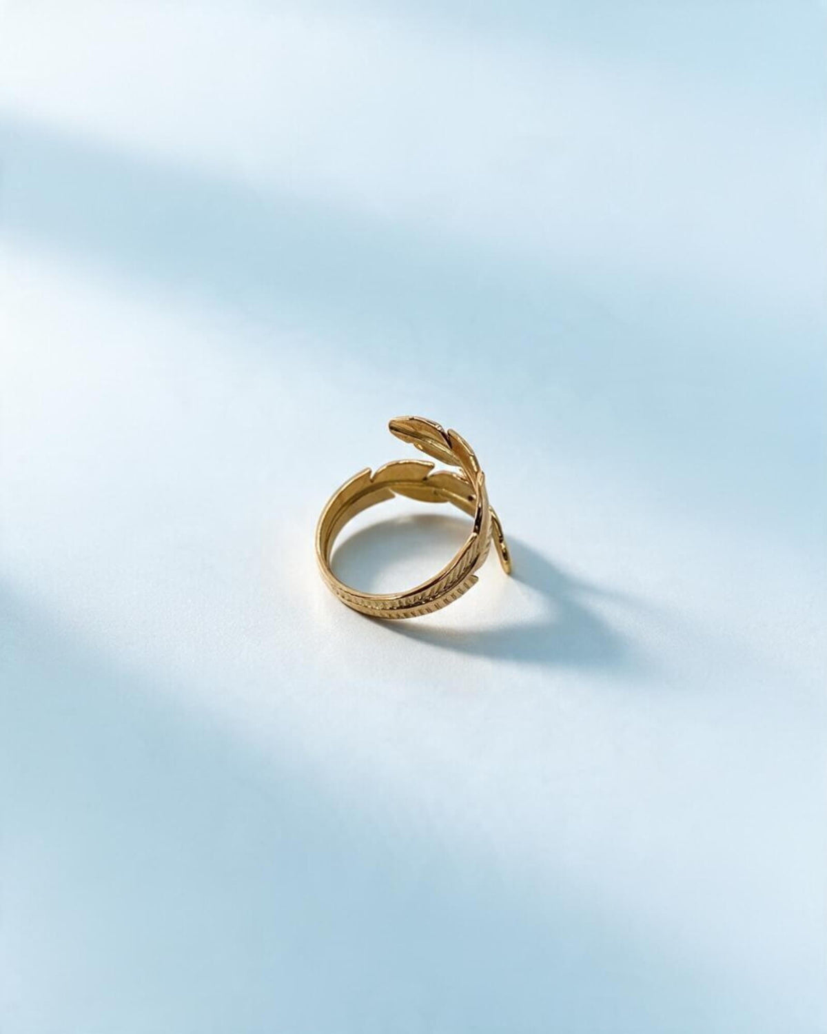 Perfil do anel Pluma dourado ajustável em aço inoxidável, mostrando a curva do aro e uma das penas levantada.