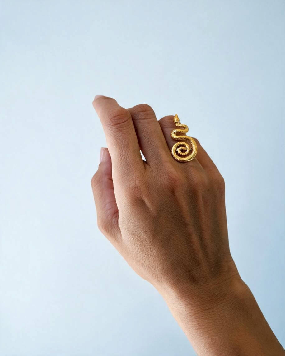 Anel serpente dourado no dedo anelar, com a mão ligeiramente fechada, destacando o corpo texturado e a cauda em espiral.