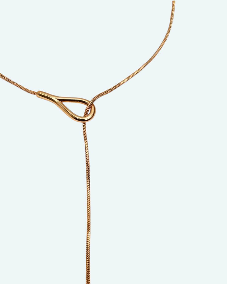 Vista detalhe zona do pecoço do colar Flow que é um cordão metálico dourado ajustável em aço inoxidável, com um design em Y que cria uma linha fluida escultural.
Contorna o pescoço, passa pela peça central e termina num pendente em forma de gota. É possível usa-lo junto ao pescoço como choker com o cordão pendurado mais comprido, ou deixa-lo mais solto para uma vibe descontraída.
Um colar para quebrar a simplicidade de uma gola redonda, para intensificar um decote em V ou contrariar um top de ombro a ombro.