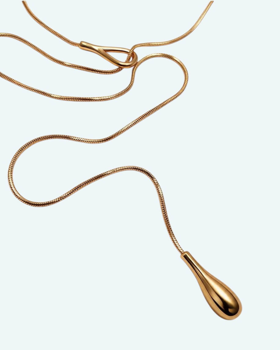 Vista detalhe zona inferior do colar Flow que é um cordão metálico dourado ajustável em aço inoxidável, com um design em Y que cria uma linha fluida escultural.
Contorna o pescoço, passa pela peça central e termina num pendente em forma de gota. É possível usa-lo junto ao pescoço como choker com o cordão pendurado mais comprido, ou deixa-lo mais solto para uma vibe descontraída.
Um colar para quebrar a simplicidade de uma gola redonda, para intensificar um decote em V ou contrariar um top de ombro a ombro.