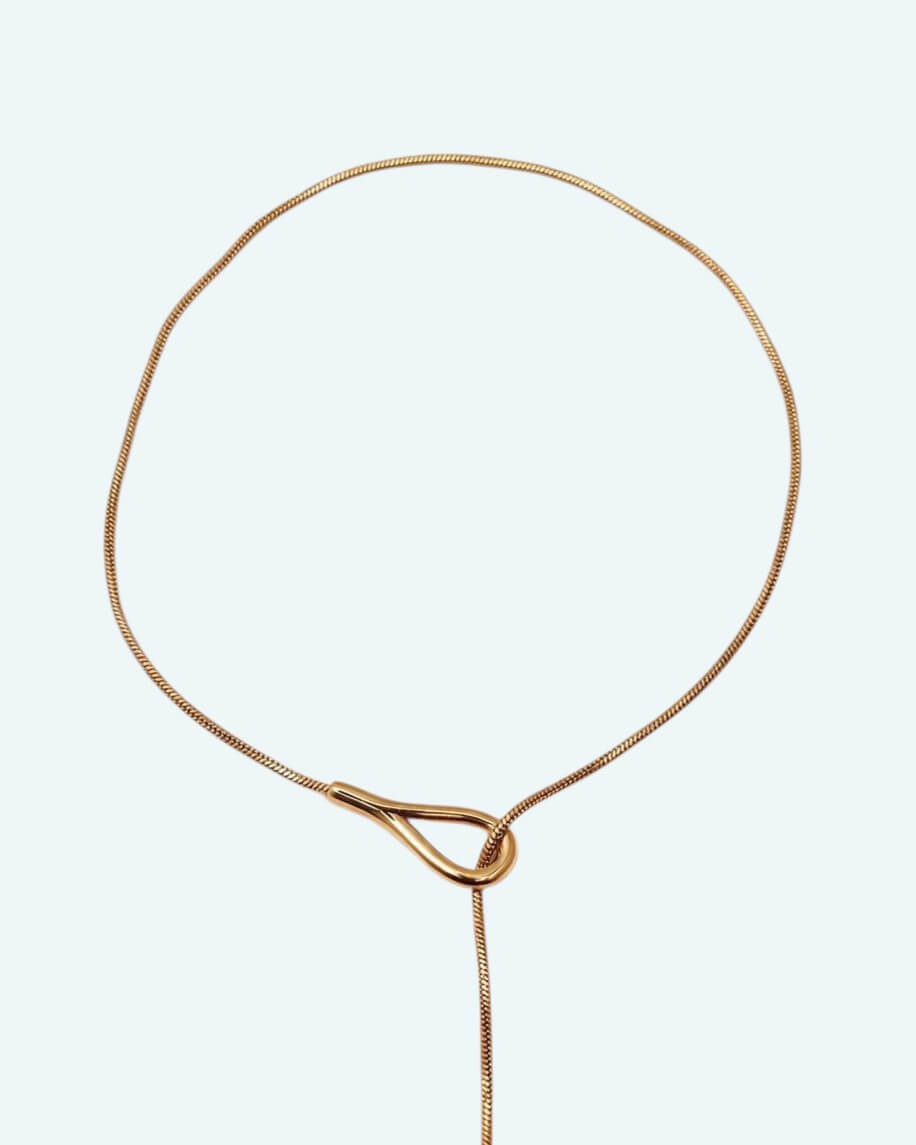 Vista detalhe zona superior do colar Flow que é um cordão metálico dourado ajustável em aço inoxidável, com um design em Y que cria uma linha fluida escultural.
Contorna o pescoço, passa pela peça central e termina num pendente em forma de gota. É possível usa-lo junto ao pescoço como choker com o cordão pendurado mais comprido, ou deixa-lo mais solto para uma vibe descontraída.
Um colar para quebrar a simplicidade de uma gola redonda, para intensificar um decote em V ou contrariar um top de ombro a ombro.