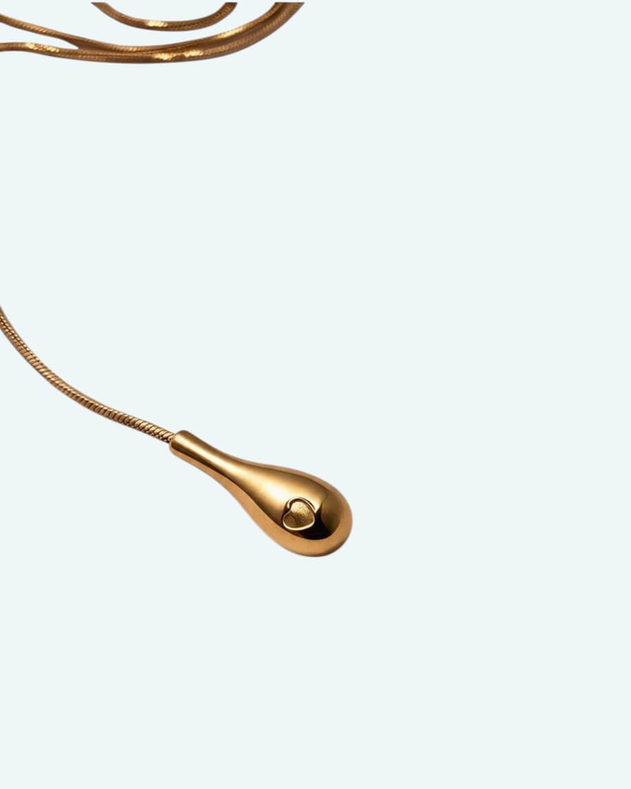 Vista detalhe do pendente do colar Flow que é um cordão metálico dourado ajustável em aço inoxidável, com um design em Y que cria uma linha fluida escultural.
Contorna o pescoço, passa pela peça central e termina num pendente em forma de gota. É possível usa-lo junto ao pescoço como choker com o cordão pendurado mais comprido, ou deixa-lo mais solto para uma vibe descontraída.
Um colar para quebrar a simplicidade de uma gola redonda, para intensificar um decote em V ou contrariar um top de ombro a ombro.