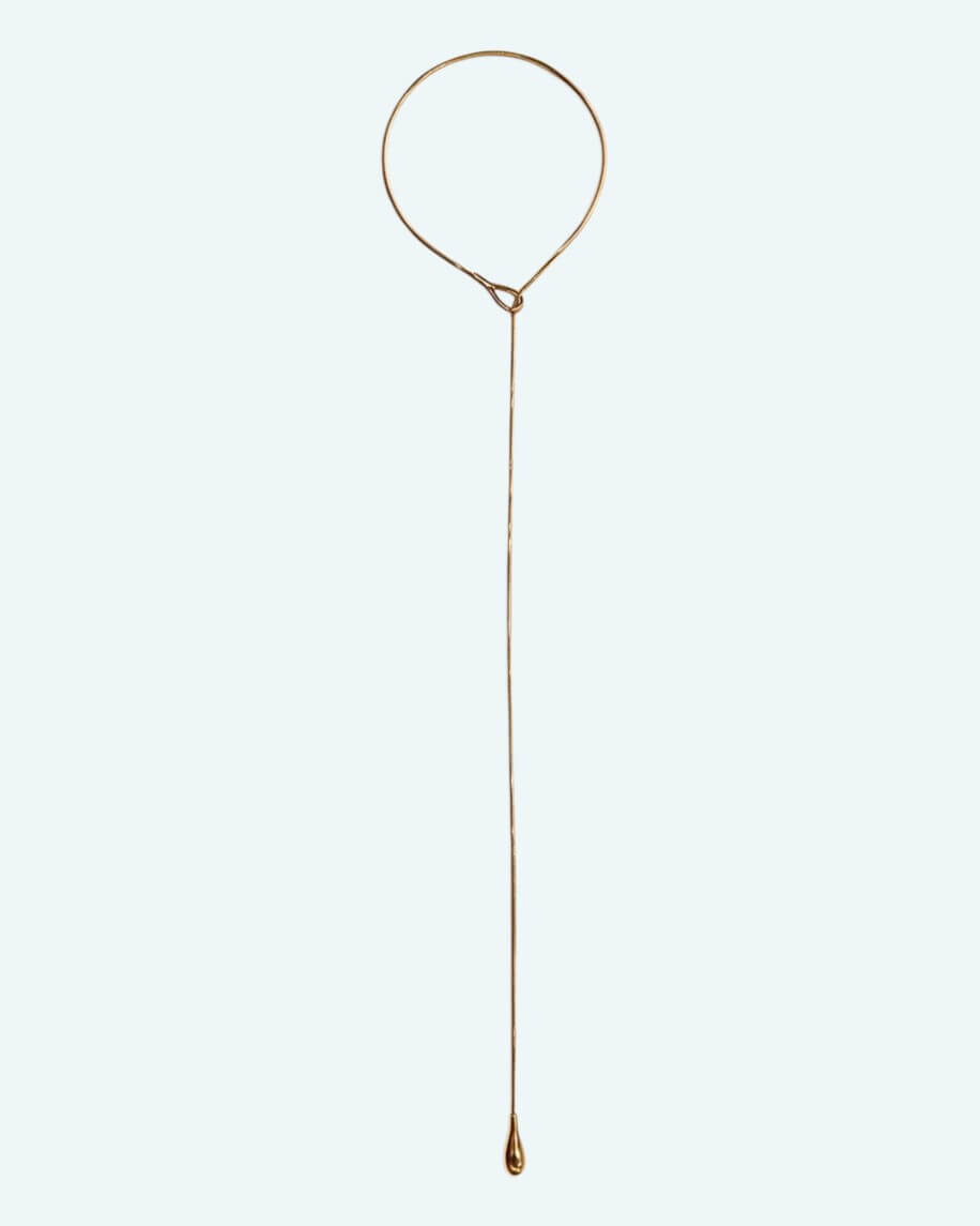Vista total e frontal do colar Flow. O Flow é um cordão metálico dourado ajustável em aço inoxidável, com um design em Y que cria uma linha fluida escultural.
Contorna o pescoço, passa pela peça central e termina num pendente em forma de gota. É possível usa-lo junto ao pescoço como choker com o cordão pendurado mais comprido, ou deixa-lo mais solto para uma vibe descontraída.
Um colar para quebrar a simplicidade de uma gola redonda, para intensificar um decote em V ou contrariar um top de ombro a ombro.