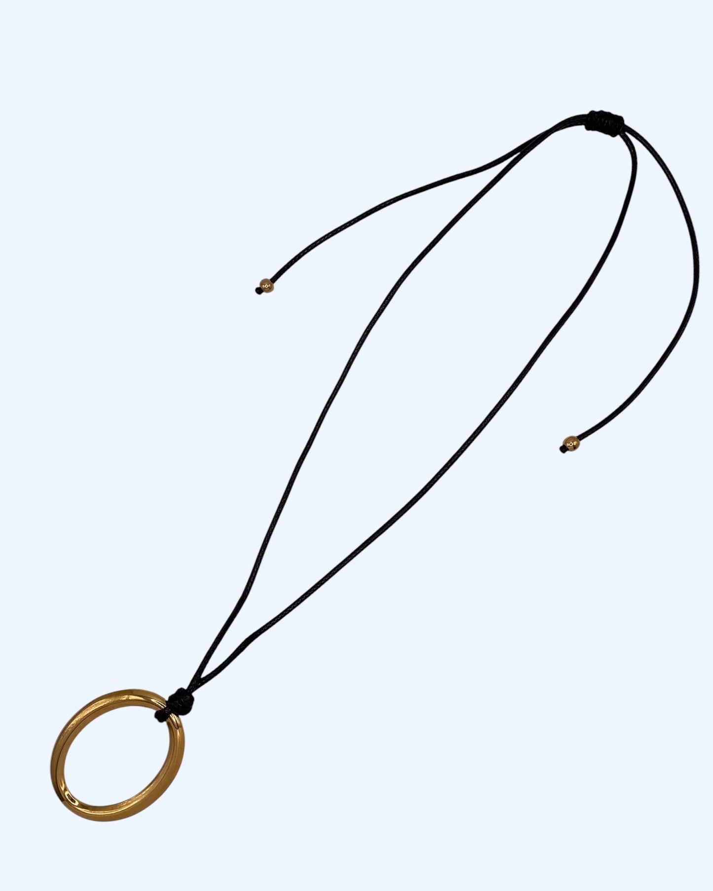 Colar comprido com medalhão oval dourado em aço inoxidável e cordão preto ajustável, fotografado em perspetiva lateral sobre fundo neutro. Colar versátil, contemporâneo e minimalista.