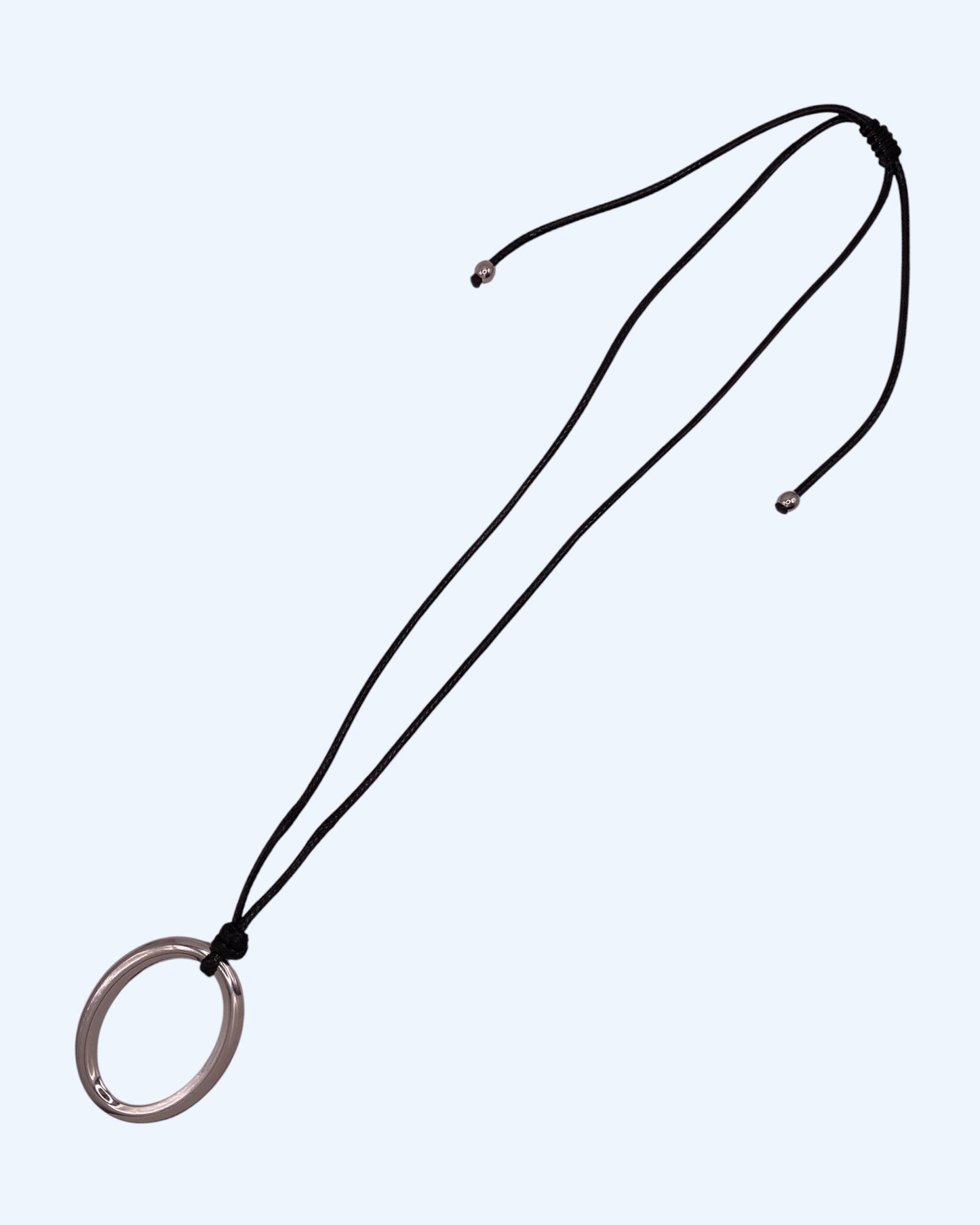 Colar comprido com medalhão oval prateado em aço inoxidável e cordão preto ajustável, fotografado em perspetiva lateral sobre fundo neutro. Colar versátil, contemporâneo e minimalista.