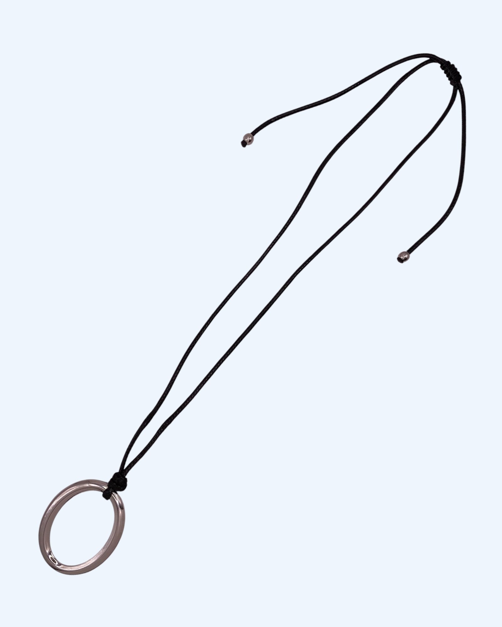 Colar comprido com medalhão oval prateado em aço inoxidável e cordão preto ajustável, fotografado em perspetiva lateral sobre fundo neutro. Colar versátil, contemporâneo e minimalista.