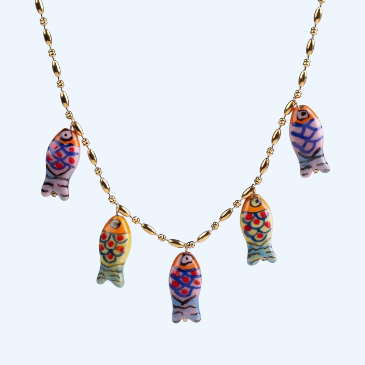 Vista em detalhe do Colar dourado em aço inoxidável e pendentes em forma de peixe coloridos em porcelana. Este colar tem um acabamento artesanal e vibe mediterrânica, é um colar divertido, leve, descontraído e um toque summer, beachy e artsy. Colar ideal para férias.