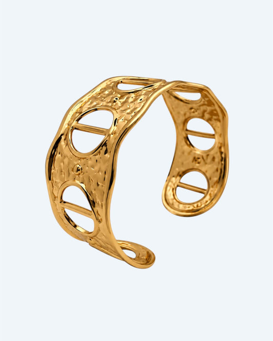 Vista na diagonal da Pulseira dourada rígida, ajustável, em aço inoxidável, martelada com aberturas. Esta pulseira é uma versão contemporânea de boho vintage. É para quem gosta de looks marcantes ou para quem procura uma pulseira para usar numa ocasião especial ou cerimónia.
