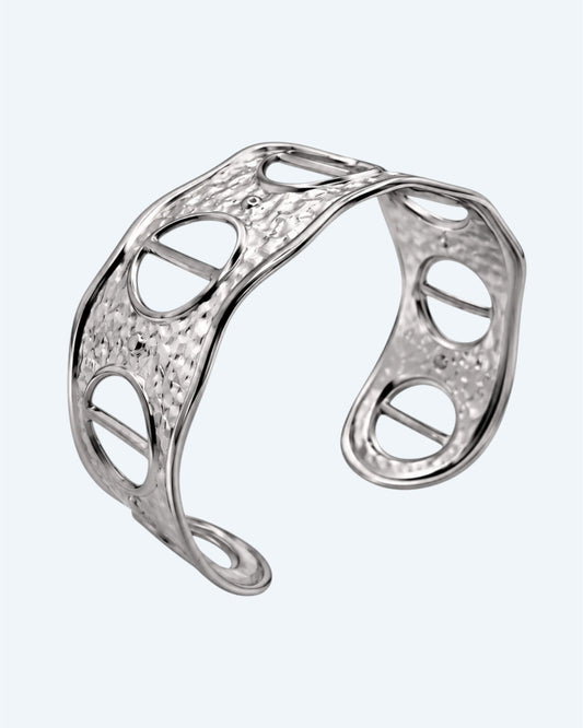 Vista na diagonal da Pulseira prateada rígida, ajustável, em aço inoxidável, martelada com aberturas. Esta pulseira é uma versão contemporânea de boho vintage. É para quem gosta de looks marcantes ou para quem procura uma pulseira para usar numa ocasião especial ou cerimónia.